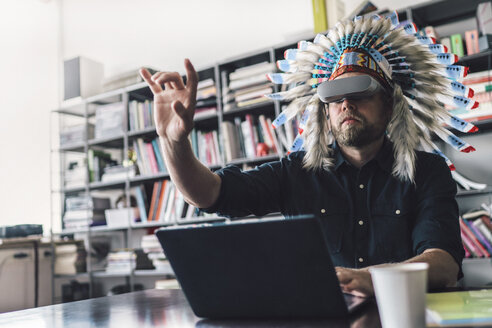 Mann mit indianischem Kopfschmuck und VR-Brille im Büro, der einen Laptop benutzt - RIBF00828