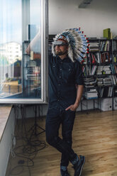 Man wearing Indian headdress and VR glasses in office, looking out of window - RIBF00830