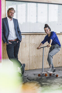 Älterer Mann und seine Assistentin spielen mit Roller, stehend im Büro vor einer weißen Tafel - RIBF00839