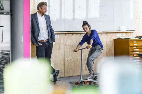 Älterer Mann und seine Assistentin spielen mit Roller, stehend im Büro vor einer weißen Tafel - RIBF00841