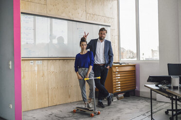 Älterer Mann und seine Assistentin spielen mit Roller, stehend im Büro vor einer weißen Tafel - RIBF00845
