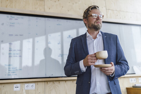 Geschäftsmann steht in seinem Büro vor einem Whiteboard und trinkt Kaffee aus einer Holztasse - RIBF00846
