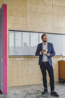 Businessman standing in his office in front of whiteboard, drinking coffee from a wooden cup - RIBF00847