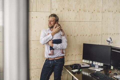 Ein berufstätiger Vater steht im Büro und hält seinen Sohn im Arm, lizenzfreies Stockfoto