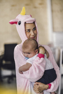 Junge Mutter mit Einhorn-Strampler, stehend im Büro, hält ihren Sohn im Arm - RIBF00862