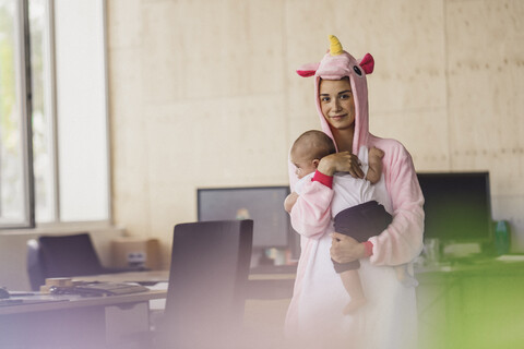 Junge Mutter mit Einhorn-Strampler, stehend im Büro, hält ihren Sohn im Arm, lizenzfreies Stockfoto