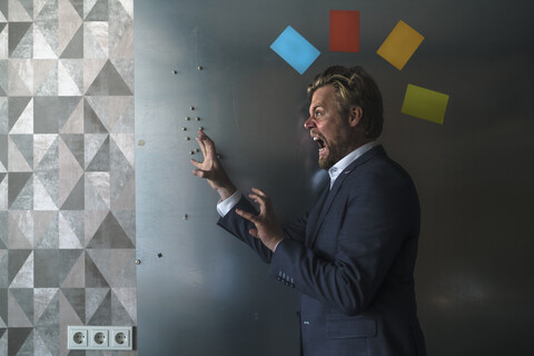Geschäftsmann, der mit einem bunten Kopfschmuck aus Haftnotizen vor einer Magnetwand steht und brüllt, lizenzfreies Stockfoto