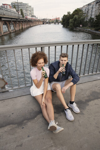 Junges Paar trinkt Bier auf einer städtischen Brücke über einen Fluss, lizenzfreies Stockfoto