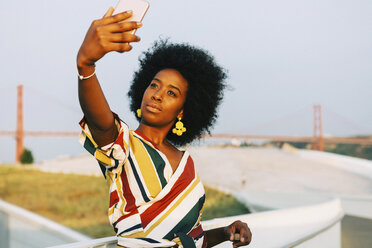 Confident young woman taking selfie with 25 de Abril Bridge in background, Lisbon, Portugal - FSIF03629