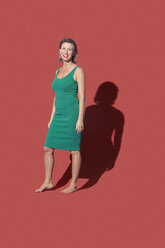 Porträt lächelnde, selbstbewusste barfüßige Frau in grünem Kleid vor rotem Hintergrund - FSIF03648