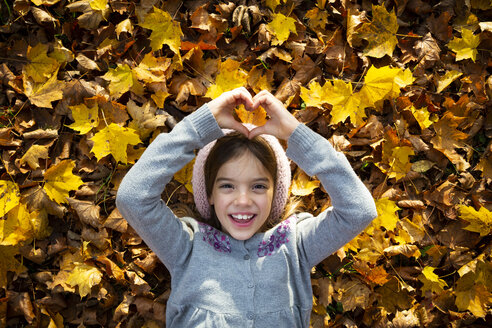 Porträt eines glücklichen kleinen Mädchens, das auf Herbstblättern liegt und mit den Händen ein Herz formt - LVF07605