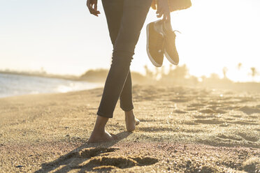 Barefoot woman on the beach, carrying her shoes - AFVF02118
