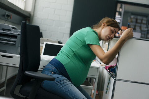 Erschöpfte schwangere Frau sitzt am Schreibtisch im Büro, lizenzfreies Stockfoto