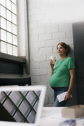 Smiling pregnant woman with disposable cup leaning against a wall in office - KNSF05449