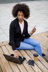 Businesswoman sitting on table outdoors using laptop and smartphone - MAUF01979