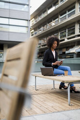 Businesswoman with laptop and coffee to go sitting on terrace looking at smartphone - MAUF01982