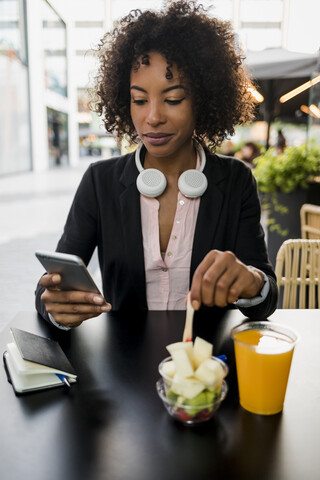Porträt einer Geschäftsfrau, die in einem Straßencafé auf ihr Handy schaut, während sie einen Obstsalat isst, lizenzfreies Stockfoto