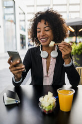 Portrait of smiling businesswoman looking at cell phone at pavement cafe while eating fruit salad - MAUF02022