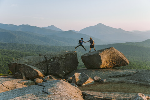 Mann hilft Frau beim Sprung über einen Felsblock, Pitchoff Mountain, Adirondack Mountains, New York State, USA, lizenzfreies Stockfoto