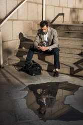 Geschäftsmann mit Tasche, der auf einer Treppe sitzt und ein Mobiltelefon benutzt - MAUF02034