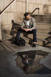 Geschäftsmann mit Tasche, der auf einer Treppe sitzt und ein Mobiltelefon benutzt - MAUF02035
