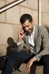 Lächelnder Geschäftsmann mit Tasche, der auf einer Treppe sitzt und mit einem Handy telefoniert - MAUF02037
