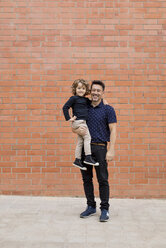 Porträt eines lächelnden Vaters, der seinen Sohn an einer Backsteinmauer trägt - MAUF02090