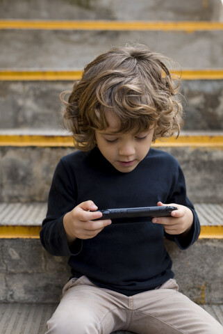 Junge sitzt auf einer Treppe und spielt mit einer Handheld-Konsole, lizenzfreies Stockfoto