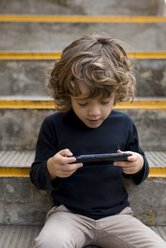 Boy sitting on stairs playing with handheld game console - MAUF02102