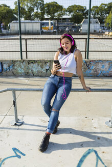 Lächelnde junge Frau mit Kopfhörern und Mobiltelefon in der Stadt - MGIF00277