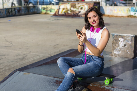 Porträt einer lächelnden jungen Frau mit Kopfhörern und Mobiltelefon in einem Skatepark, lizenzfreies Stockfoto