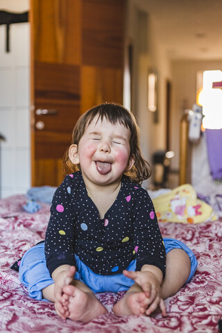 Porträt eines Kleinkindes, das barfuß auf dem Bett sitzt und die Zunge herausstreckt, lizenzfreies Stockfoto