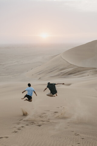 Namibia, Namib, Rückenansicht von zwei Freunden, die auf einer Wüstendüne in die Luft springen, lizenzfreies Stockfoto