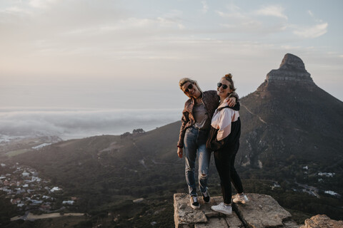 Südafrika, Kapstadt, Kloof Nek, Porträt von zwei glücklichen Frauen, die bei Sonnenuntergang auf einem Felsen stehen, lizenzfreies Stockfoto