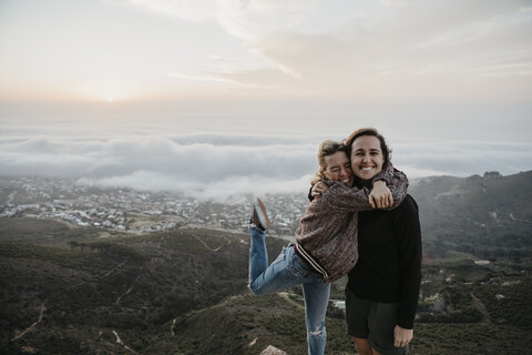 Südafrika, Kapstadt, Kloof Nek, Porträt von zwei glücklichen Frauen, die sich bei Sonnenuntergang umarmen, lizenzfreies Stockfoto