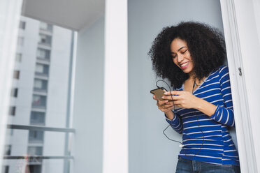 Portrait of smiling young woman using smartphone and earphones at home - KKAF03073