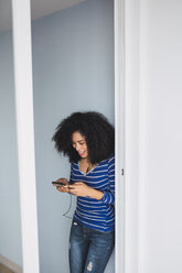 Smiling young woman using smartphone and earphones at opened balcony door - KKAF03074