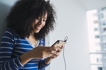 Porträt einer lächelnden jungen Frau mit Kopfhörern, die auf ihr Mobiltelefon schaut - KKAF03076