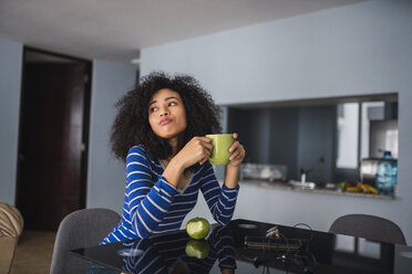 Portrait of young woman drinking coffee and eating an apple at home - KKAF03081