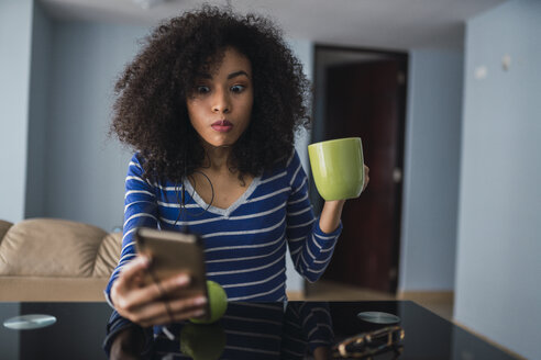 Porträt einer jungen Frau mit Tasse, die auf ein Mobiltelefon starrt - KKAF03084