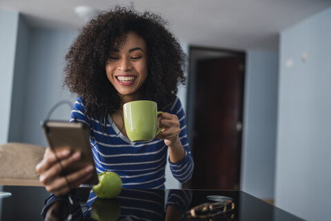 Porträt einer glücklichen jungen Frau mit Kaffeetasse, die auf ihr Handy schaut, lizenzfreies Stockfoto
