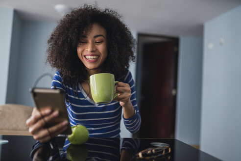 Porträt einer glücklichen jungen Frau mit Kaffeetasse, die auf ihr Handy schaut - KKAF03085