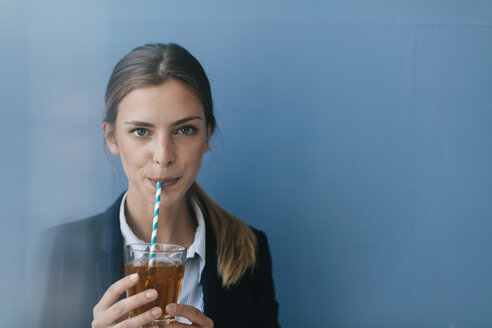 Porträt einer jungen Geschäftsfrau vor blauem Hintergrund, die Eistee mit einem Strohhalm trinkt - GUSF01691