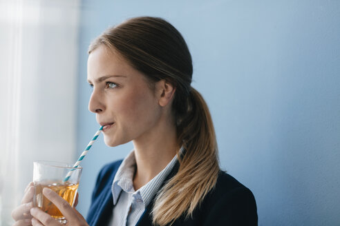 Porträt einer jungen Geschäftsfrau vor blauem Hintergrund, die Eistee mit einem Strohhalm trinkt - GUSF01692