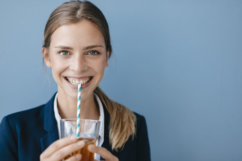 Porträt einer jungen Geschäftsfrau vor blauem Hintergrund, die Eistee mit einem Strohhalm trinkt - GUSF01693