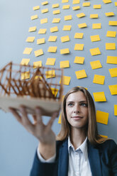 Junge Geschäftsfrau hält Architekturmodell mit gelben Haftnotizen an der Wand hinter sich - GUSF01716