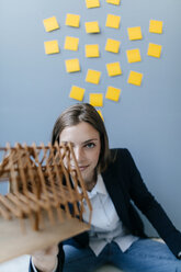 Junge Geschäftsfrau hält Architekturmodell mit gelben Haftnotizen an der Wand hinter sich - GUSF01721