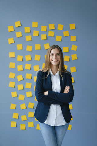 Yong Geschäftsfrau steht vor einer Wand, voll mit gelben Haftnotizen, mit verschränkten Armen, lizenzfreies Stockfoto