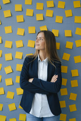 Yong businesswoman standing in front of wall, full of yellow sticky notes, with arms crossed - GUSF01779