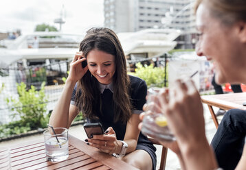 Zwei glückliche Frauen mit Smartphone und Getränken auf einer Terrasse - MGOF03890
